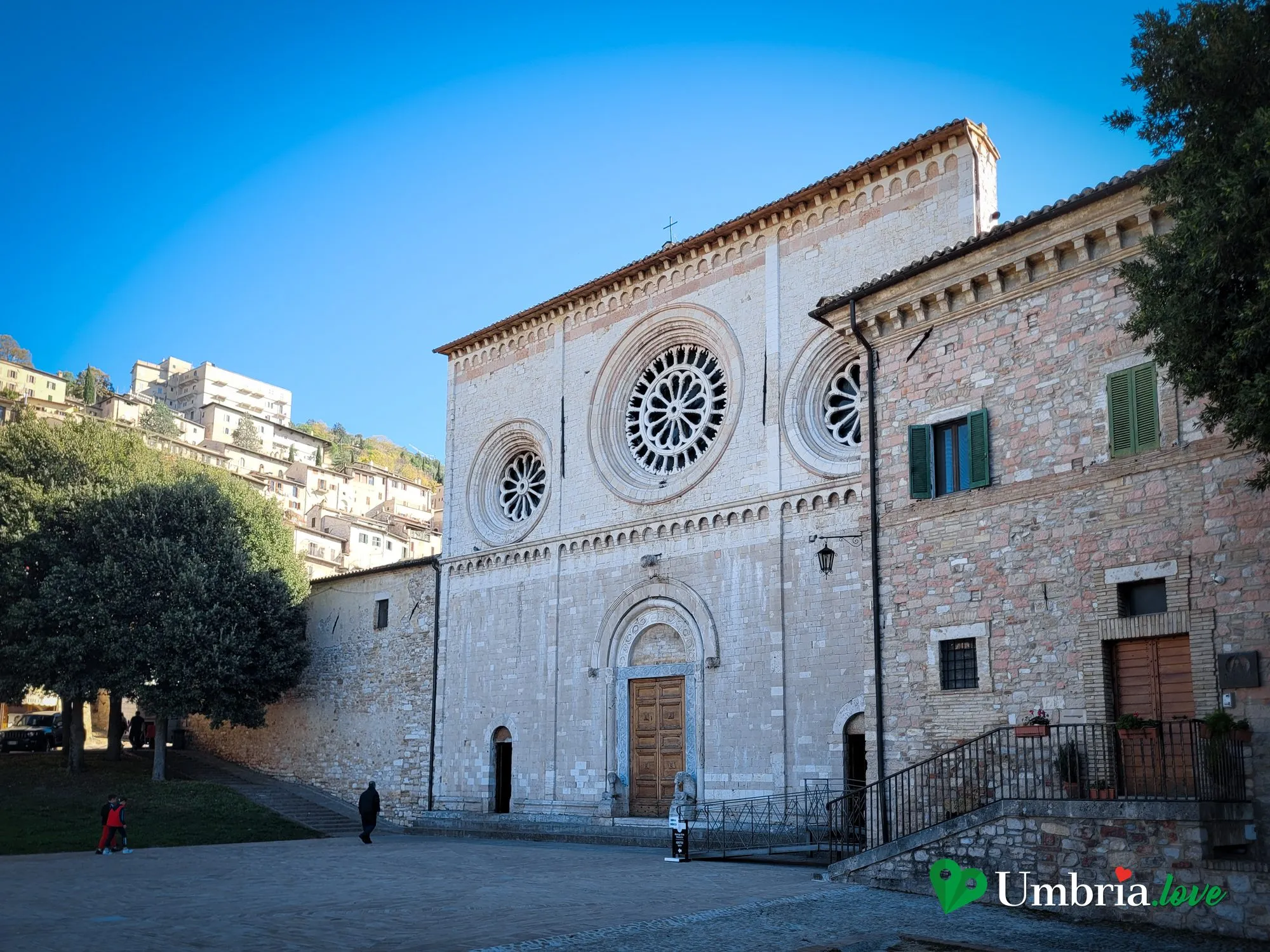 La facciata romanica dell'abbazia di San Pietro ad Assisi, con i tre rosoni e il coronamento orizzontale, sul prato che guarda la valle