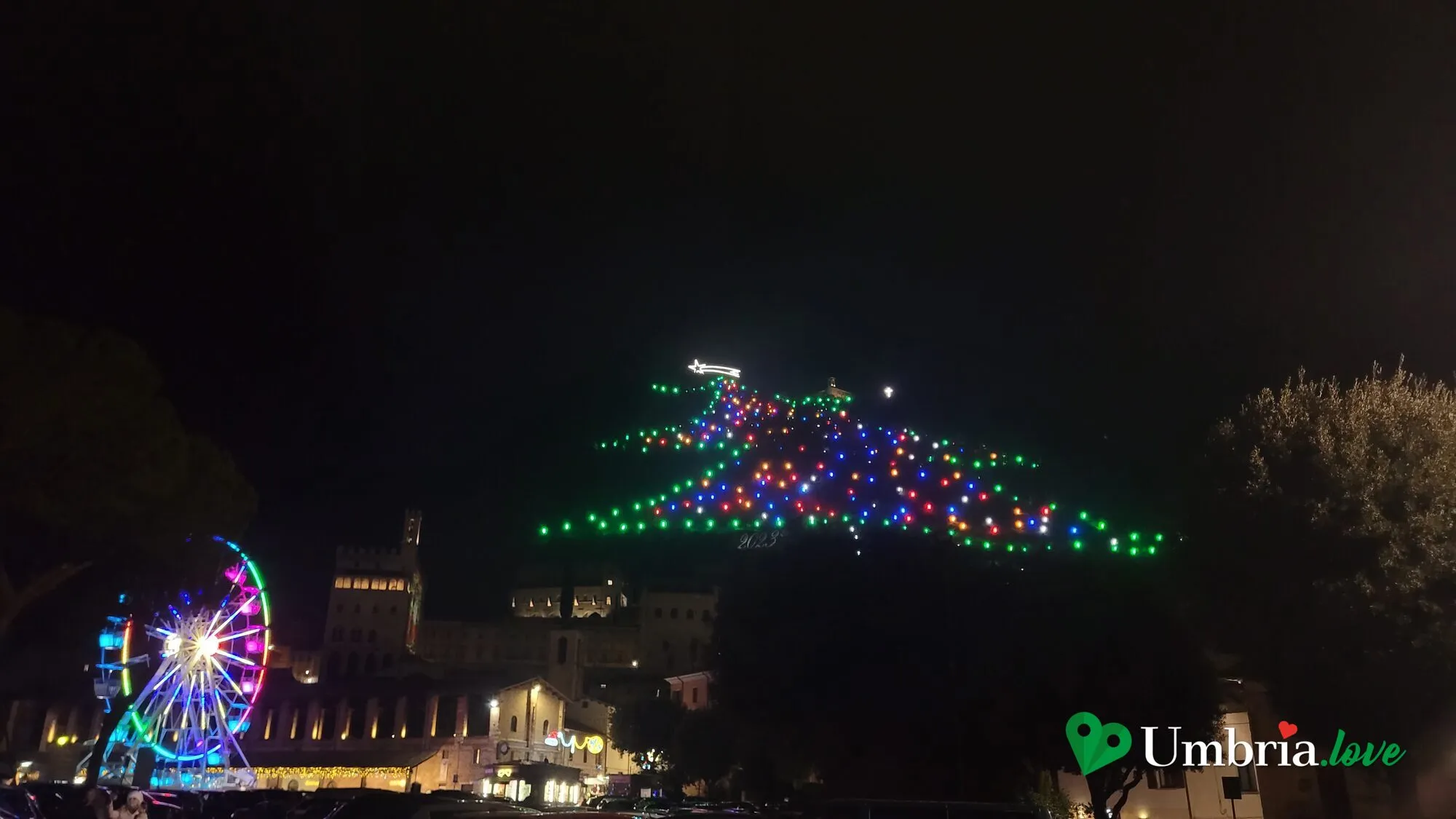 L'Albero di Natale di Gubbio: le luci disposte a forma di abete sul fianco del Monte Ingino nella notte, con la stella cometa in cima; in primo piano la ruota panoramica e il Palazzo dei Consoli illuminati