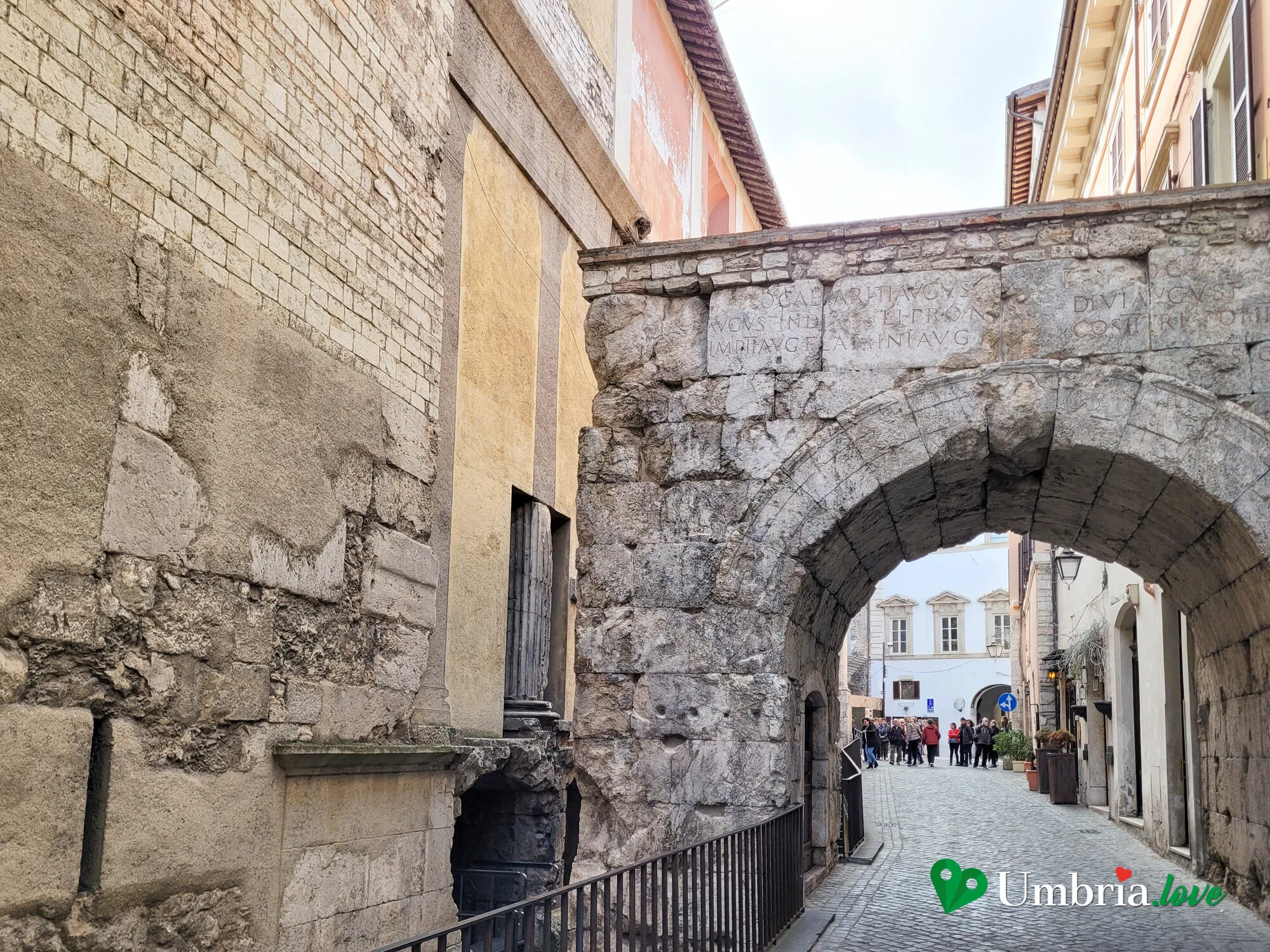 L'Arco di Druso e Germanico a Spoleto, con la chiesa di Sant'Ansano sullo sfondo e la gente che passa sotto l'arco