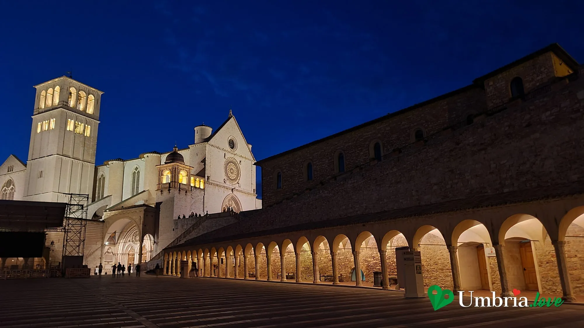 La Basilica di San Francesco di Assisi illuminata al crepuscolo, con il loggiato ad archi della piazza inferiore in primo piano