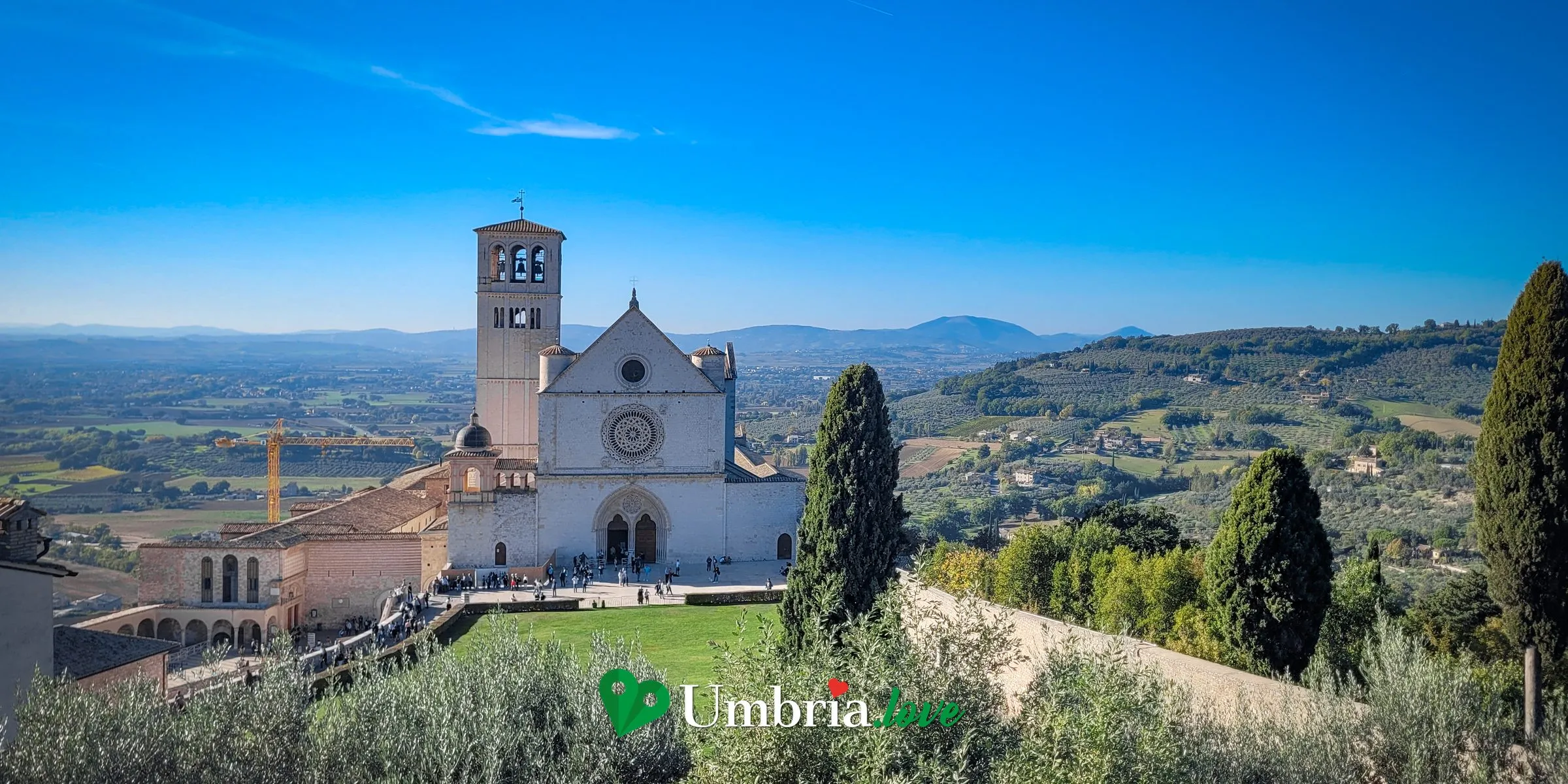 La Basilica di San Francesco di Assisi vista dall'alto, dalla parte alta della città: la facciata gotica, il campanile romanico e il lungo Sacro Convento sulle arcate, con la Valle Umbra e gli uliveti che si aprono dietro