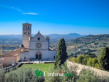La Basilica di San Francesco di Assisi vista dall'alto, dalla parte alta della città: la facciata gotica, il campanile romanico e il lungo Sacro Convento sulle arcate, con la Valle Umbra e gli uliveti che si aprono dietro