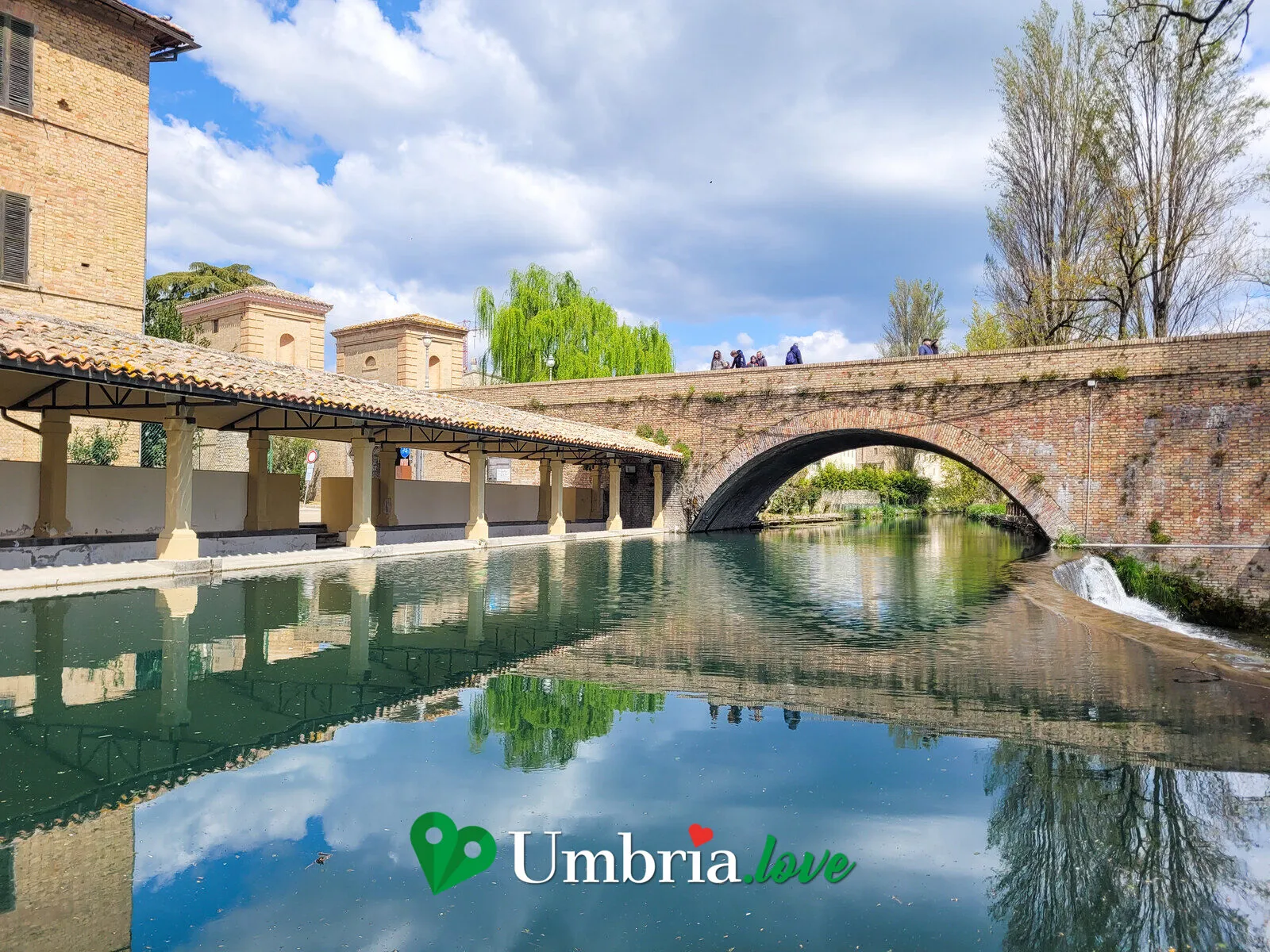 Il portico medievale del Lavatoio dell'Accolta a Bevagna, con un ponte a schiena d'asino riflesso nell'acqua verde