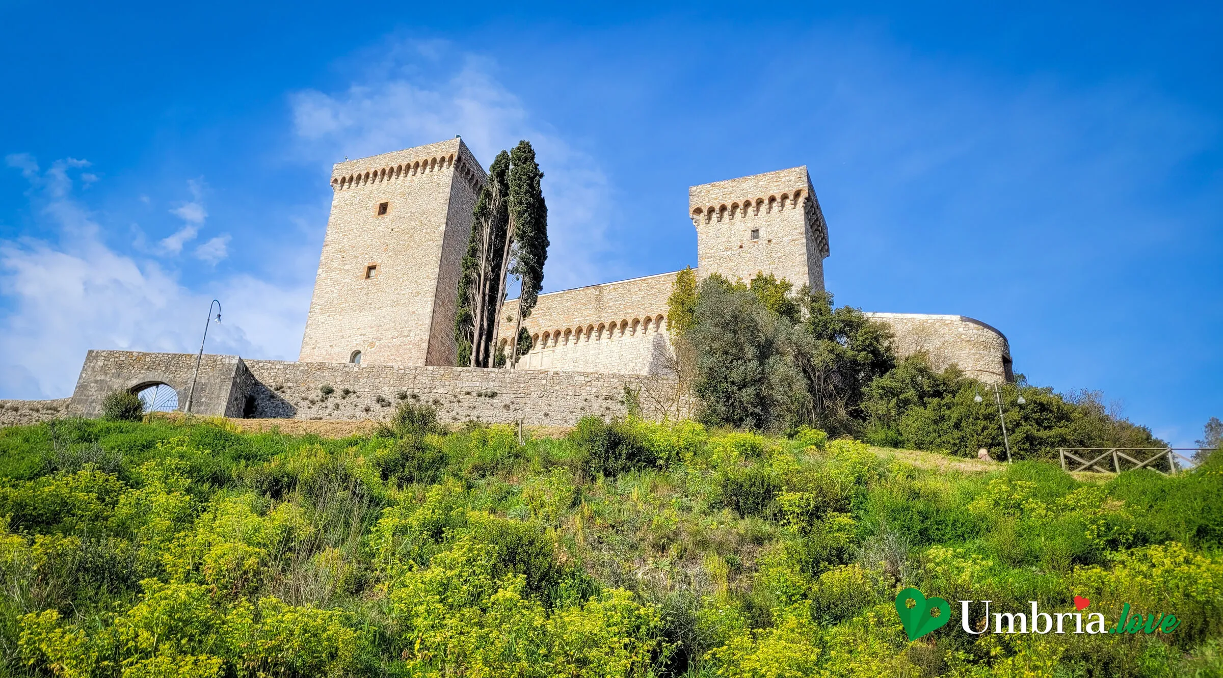 La Rocca Albornoz di Narni, con i due torrioni merlati e un cipresso, tra i fiori di primavera sul colle