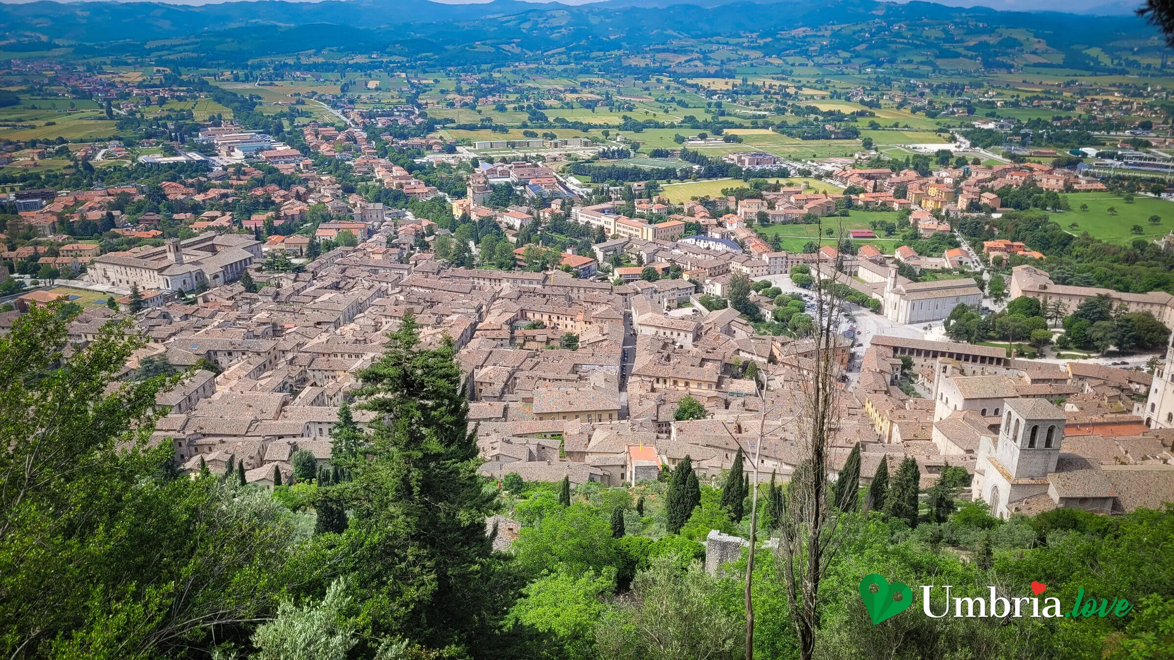 Gubbio vista dall'alto: il centro storico di pietra grigia che sale in file di tetti sul fianco del Monte Ingino, con la torre del Palazzo dei Consoli al centro e la pianura eugubina sullo sfondo