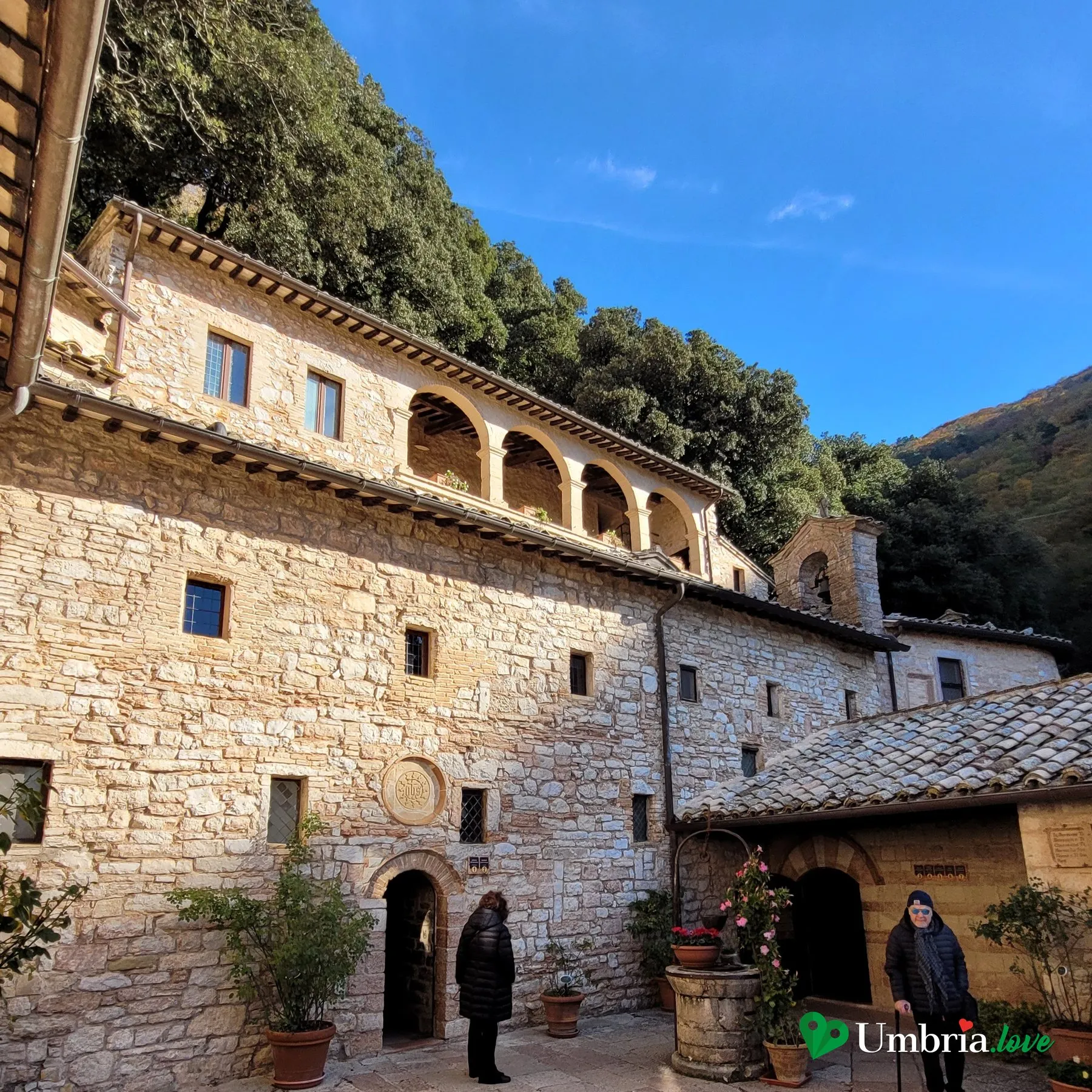 Il cortile dell'Eremo delle Carceri di Assisi: gli edifici di pietra con la loggia e il campanile a vela addossati alla lecceta del Monte Subasio, il pozzo e due visitatori