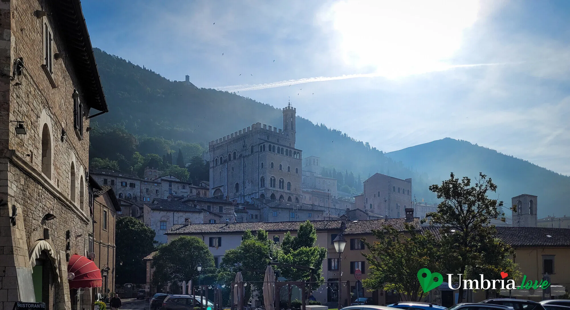 Gubbio vista dal basso al mattino: il Palazzo dei Consoli e il Palazzo del Podestà emergono dai tetti, con il Monte Ingino alle spalle avvolto nella foschia