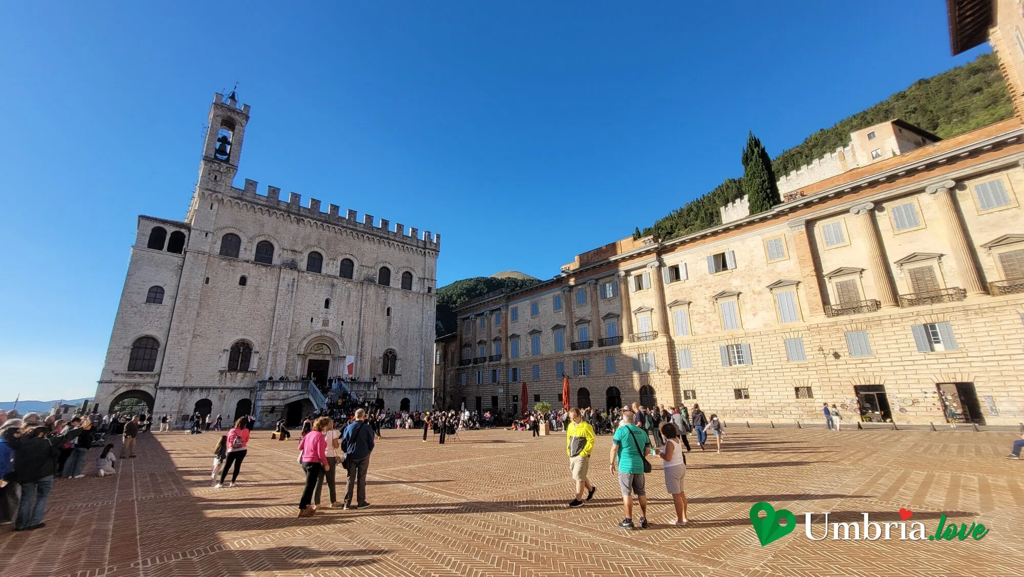 Piazza Grande a Gubbio: l'ampio spiazzo pavimentato in cotto della piazza pensile, con il Palazzo dei Consoli e la sua torre campanaria a sinistra, il palazzo Ranghiasci sulla destra e il Monte Ingino alle spalle