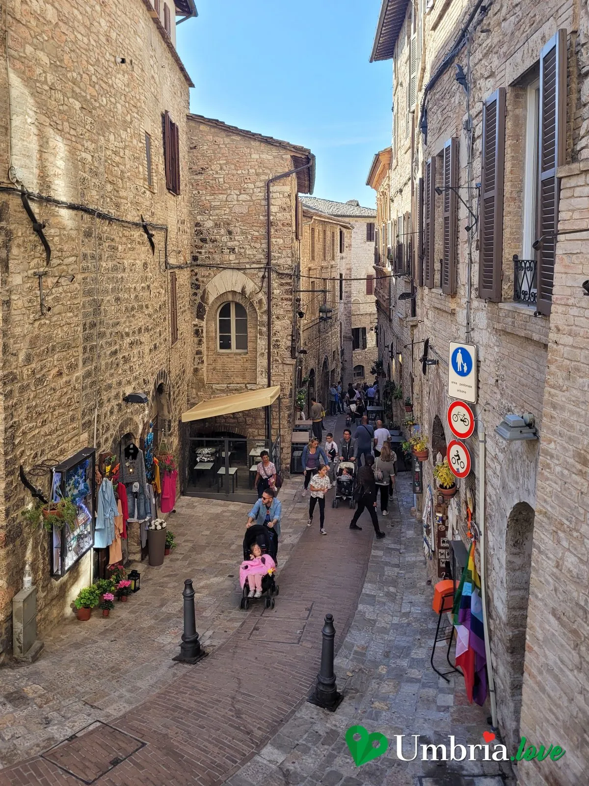 Una via del centro di Assisi in salita, tra le case di pietra chiara del Subasio, con le insegne delle botteghe